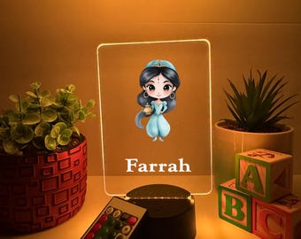Könnte beinhalten: Ein dekoratives LED-Nachtlicht mit einer Cartoon-Prinzessin in einem blauen Kleid, mit dem Namen "Farrah" darunter gedruckt. Das Licht steht auf einem schwarzen Sockel, und eine Fernbedienung ist im Lieferumfang enthalten. Der Hintergrund umfasst Topfpflanzen und ABC-Blöcke.