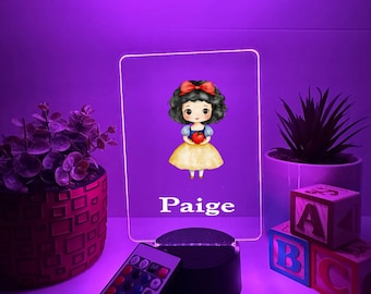 Könnte beinhalten: LED-Nachtlicht mit Schneewittchen-Motiv und dem Namen "Paige". Die Acrylplatte zeigt eine Cartoon-Illustration von Schneewittchen mit einem Apfel. Eine Fernbedienung und Buchstabenblöcke sind ebenfalls vorhanden.
