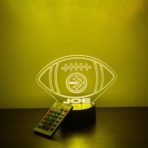 May include: A glowing yellow football-shaped LED lamp with the word "JOE" illuminated at the base. The lamp features the Pittsburgh Steelers logo in the center. A remote control with colorful buttons sits to the left.