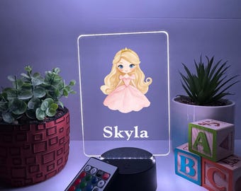 Könnte beinhalten: Ein dekoratives LED-Nachtlicht mit einer Prinzessinnen-Illustration in einem rosa Kleid und dem Namen "Skyla". Das Licht steht auf einem schwarzen Sockel mit einer Fernbedienung. Dekorative Pflanzen und ABC-Blöcke befinden sich im Hintergrund.