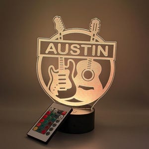 May include: A circular acrylic LED lamp featuring the name AUSTIN in a horizontal bar, with silhouettes of an electric guitar and an acoustic guitar behind it. The lamp is mounted on a black cylindrical base and includes a small remote control.