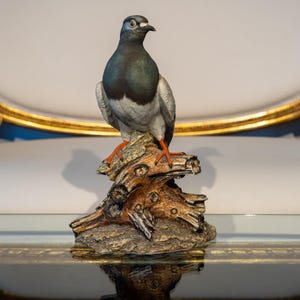 May include: A ceramic figurine of a gray and white pigeon perched on a brown tree branch. The bird has orange feet and is standing on a textured base.