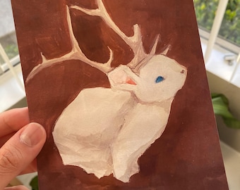May include: A watercolor painting of a white rabbit with antlers on a brown background. The rabbit has blue eyes and pink ears. The artwork is on a rectangular piece of paper.