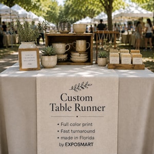 May include: A beige fabric table runner featuring black text that reads Custom Table Runner, Full color print, Fast turnaround, made in Florida by EXPOSMART, with a leaf graphic. The runner is displayed on a table with ceramic mugs and soap.