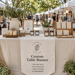 May include: A beige linen table runner featuring a central logo with a leaf illustration and the text Handcrafted With Care, Custom Table Runner, Full color print, Fast turnaround, made in Florida by EXPOSMART, displayed on a vendor table.