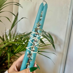 May include: Two teal blue candles are bound together with twine and herbs. The candles are long and thin, and the herbs are visible through the wax.