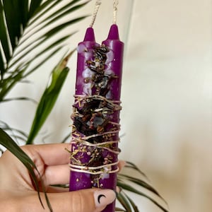 May include: Two purple ritual candles, adorned with dried herbs and twine. The candles are cylindrical with a flat top and a wick. The candles are wrapped with twine and decorated with dried flowers and herbs. The background includes green leaves.