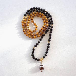 May include: A layered necklace with black and tiger's eye beads, and gold-coloured accents. The necklace has a pendant shaped like a football. The beads are strung together, creating a layered effect. The necklace is displayed on a white surface.
