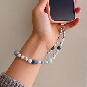 May include: A beaded phone wrist strap featuring a mix of white, blue, and gray beads. The strap is attached to a pink phone case with a gold-colored clasp. The beads are strung together to create a stylish accessory.