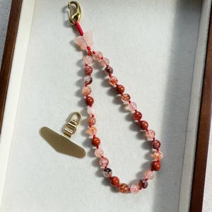 May include: A beaded phone charm with a gold-colored clasp and a matching phone grip. The charm features a string of red and pink beads, with a small butterfly accent. The charm is displayed in a wooden box.