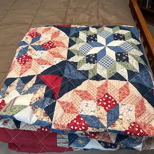 May include: A folded quilt featuring a starburst design. The quilt showcases a patchwork of red, blue, and white fabrics with floral and geometric patterns. The quilt is folded on a bed with a brown comforter.