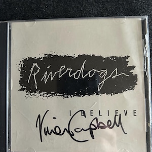 May include: A CD case with a light grey cover. The title "Riverdogs" is in white, stylized text above the words "I Believe" and a signature. The CD case is in a clear plastic sleeve.