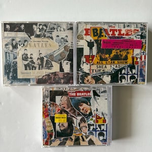 May include: Three Beatles CD albums in clear plastic cases. The album covers feature collages of images, text, and the band's name. One album has a pink sticker. The text "BEST BUY" and a price tag are visible on one album.