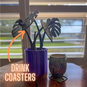 May include: Purple drink coasters with a black plant in the centre. The plant has three leaves that look like monstera leaves. The coasters are made of a 3D printed material. The text "DRINK COASTERS" is visible in the image.