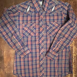 May include: A blue, red, and beige plaid long-sleeved shirt with a button-up front and two chest pockets. The shirt has a lightning bolt design on each shoulder.