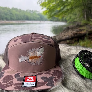 May include: A brown camouflage snapback hat with a mesh back and a flat bill. The front panel features an embroidered fly fishing lure in shades of tan, white, and orange. A black and white sticker on the bill reads Richardson Flat Bill Snapback.
