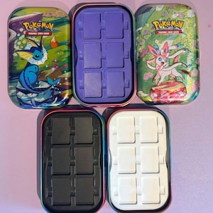 May include: Two rectangular Pokémon Trading Card Game metal tins featuring Vaporeon and Sylveon illustrations, alongside three rectangular plastic inserts in purple, black, and white, each containing six recessed slots for organising trading cards.
