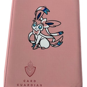 May include: Pink card binder with a cartoon Sylveon character. The binder has a stitched border and the words "CARD GUARDIAN" printed below a shield logo. The Sylveon character is white, pink, and blue.