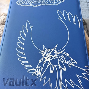 Pode incluir: Um fichário de cartas azul com o texto "Scarlet & Violet Paradox Rift" e "vaultx" em branco. Uma ilustração branca de uma criatura está na frente. O fichário é projetado para guardar cartas colecionáveis.