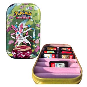 May include: A gold-coloured metal tin with a floral and Pokémon design, featuring a Sylveon character. The tin is open, revealing several game cartridges in a pink velvet-lined interior. The Pokémon Trading Card Game logo is visible.
