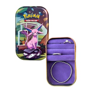 May include: A Pokemon Trading Card Game tin featuring a pink Espeon character and a purple jewelry box with a ring and bracelet. The tin has a colorful landscape design, and the jewelry box has a gold trim.