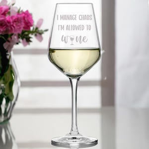 Può includere: Un bicchiere di vino in vetro trasparente con vino bianco al suo interno. Il bicchiere ha inciso il testo "I manage chaos I'm allowed to wine".