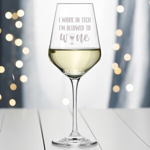 May include: A clear glass wine glass filled with white wine. The glass has a white inscription that reads "I work in tech I'm allowed to wine" with a small wine glass icon.