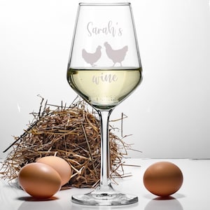 Può includere: Un bicchiere di vino in vetro trasparente con il testo "Sarah's wine" e due silhouette di galline incise sul lato. Il bicchiere è pieno di vino bianco e si trova su una superficie bianca accanto a un nido di fieno e due uova marroni.
