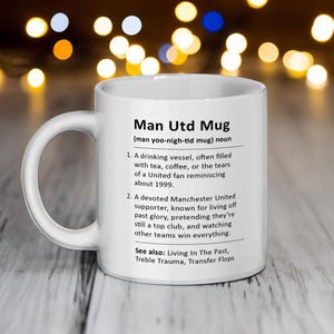 May include: White ceramic mug with a black and white definition of "Man Utd Mug" with the pronunciation "man yoo-nigh-tid mug". The definition includes two points and a "See also" section with three terms.