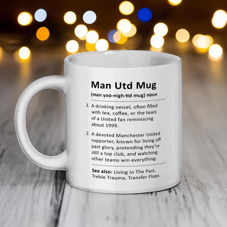 May include: White ceramic mug with a black and white definition of "Man Utd Mug" with the pronunciation "man yoo-nigh-tid mug". The definition includes two points and a "See also" section with three terms.