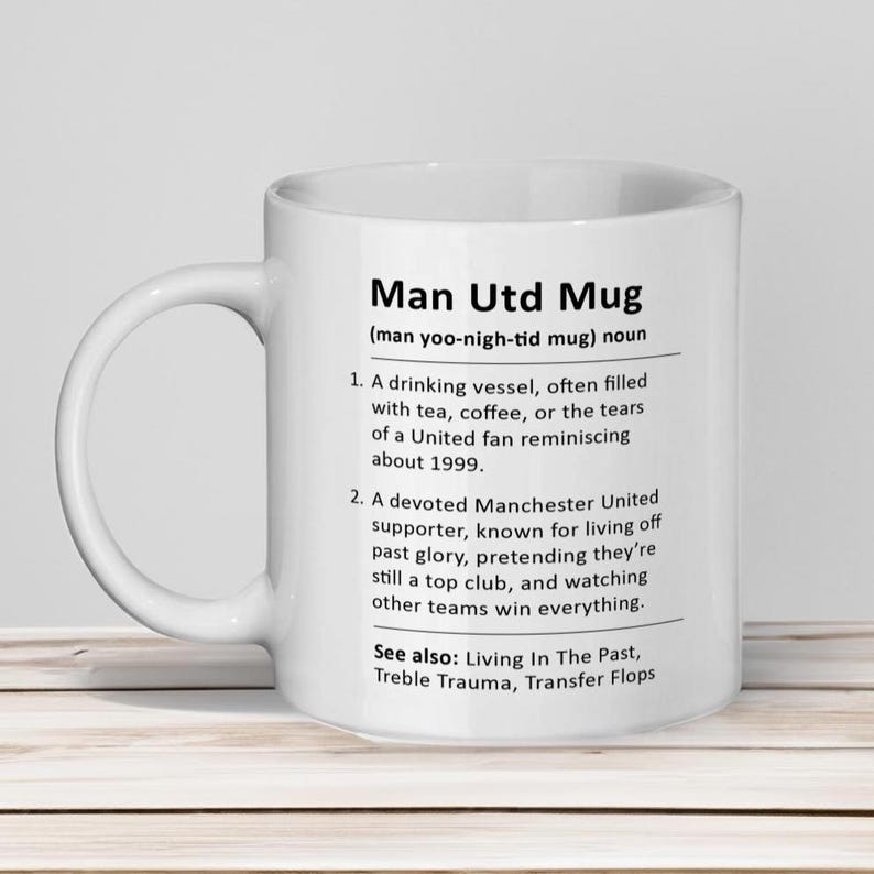 May include: White ceramic mug with a black design that defines "Man Utd Mug" as a drinking vessel often filled with tea, coffee, or tears of a United fan reminiscing about 1999. It also describes a devoted Manchester United supporter known for living off past glory, pretending they're still a top club, and watching other teams win everything. The text also includes "See also: Living In The Past, Treble Trauma, Transfer Flops."