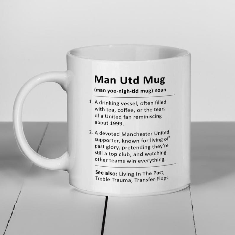 May include: White ceramic mug with a black and white design. The mug features the text "Man Utd Mug" and a definition of the term. The definition includes the text "(man yoo-nigh-tid mug) noun" and a list of characteristics.