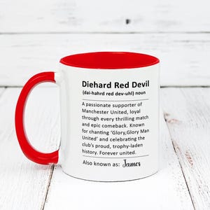 May include: White ceramic mug with a red rim and handle. The mug features a black graphic with the text "Diehard Red Devil (dai-hahrd red dev-uhl) noun" and a definition of the term. The text also includes the name "James".