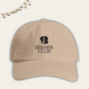 May include: A beige baseball cap with a black embroidered image of a Bernese Mountain Dog and the text "BERNER CLUB".