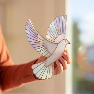 Puede incluir: Una paloma de vidriera con las alas extendidas, con paneles de vidrio iridiscentes y blancos. El ave tiene un pico rojo y está delineada con un borde metálico. Una pieza decorativa para la decoración del hogar.
