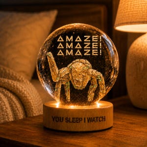 Può includere: Una sfera decorativa in vetro con un disegno di ragno e la parola "AMAZE!" ripetuta tre volte. La sfera è posizionata su una base di legno con la scritta "YOU SLEEP I WATCH". La lampada emette una calda luce.