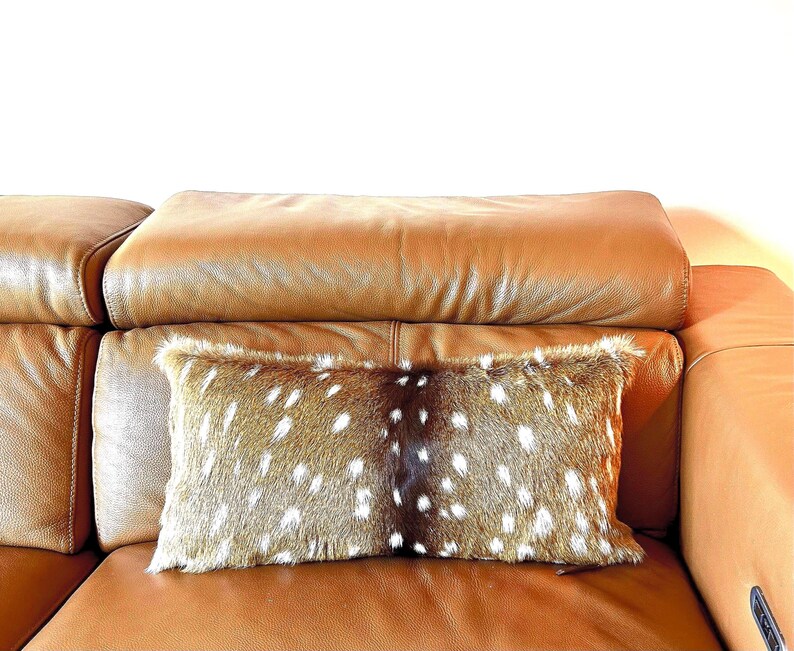 May include: A rectangular decorative pillow with a spotted pattern, resembling deer fur, rests on a brown leather sofa. The pillow has a brown and white spotted design, adding a rustic touch to the interior decor.