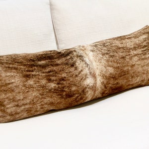 May include: A long brown and white cowhide lumbar pillow. The pillow is lying on a white couch.