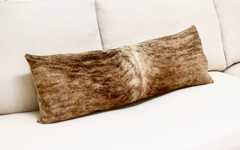 May include: A long brown and white cowhide lumbar pillow. The pillow is lying on a white couch.