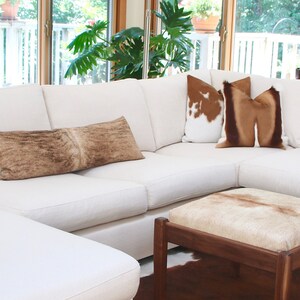 May include: A large white sectional sofa with a chaise lounge, featuring a variety of throw pillows in brown, white, and beige. The sofa is positioned in a living room with a wooden coffee table and a rug.