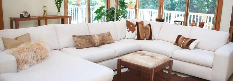 May include: A large white sectional sofa with a chaise lounge, featuring a variety of throw pillows in brown, white, and beige. The sofa is positioned in a living room with a wooden coffee table and a rug.