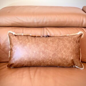 May include: A rectangular brown leather pillow with a distressed texture and a white fur trim, resting on a brown leather sofa. The pillow is a decorative accent.