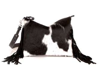 May include: A black and white cowhide shoulder bag with a black leather strap and fringe. The bag has a rectangular shape with a flap closure and a silver-tone clasp. The bag's dimensions are not visible.