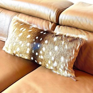 May include: A rectangular decorative pillow with a brown and white spotted pattern, resembling deer fur, rests on a tan leather sofa. The pillow has a soft, textured appearance and a small, brown leather tag.