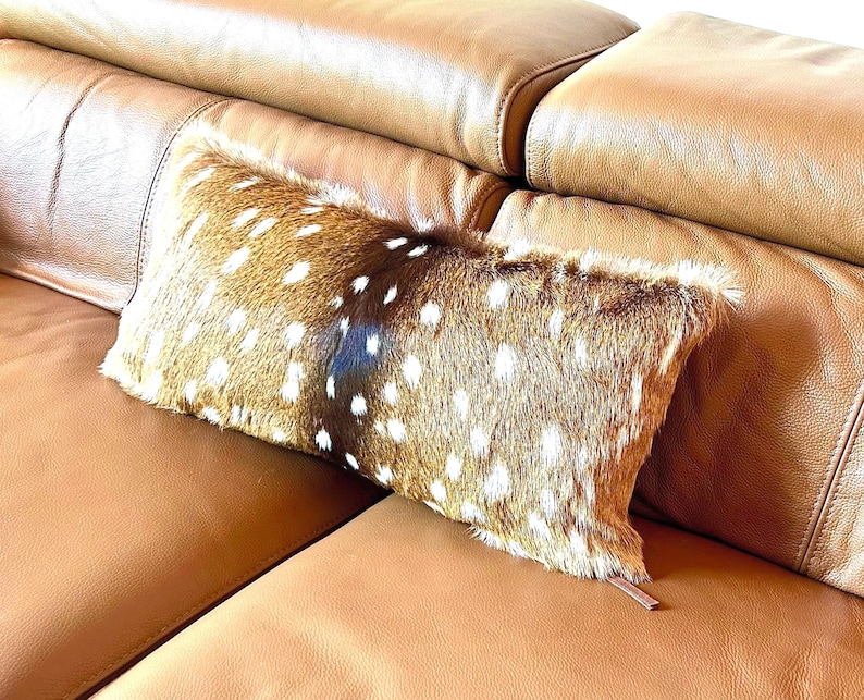 May include: A rectangular decorative pillow with a brown and white spotted pattern, resembling deer fur, rests on a tan leather sofa. The pillow has a soft, textured appearance and a small, brown leather tag.