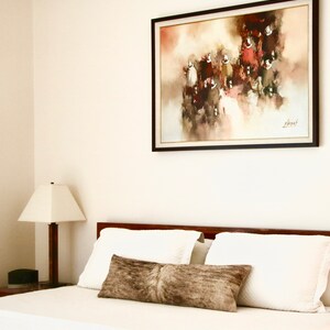 May include: A white bed with a brown headboard and white bedding. There is a brown throw pillow on the bed. A framed painting of a group of people in red and brown clothing hangs on the wall above the bed. The painting has a white background with a light brown and red abstract design.