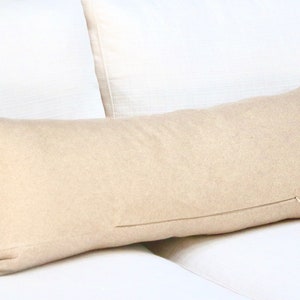 May include: A long, beige lumbar pillow with a zipper closure. The pillow is lying on a white couch.
