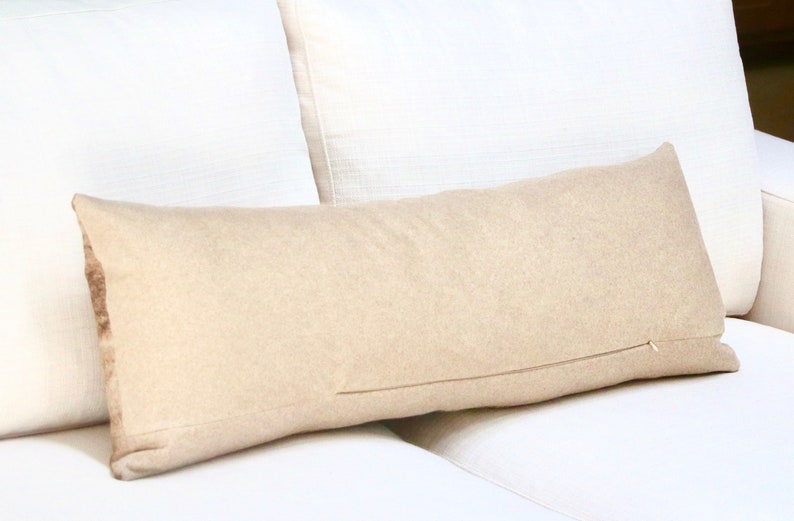 May include: A long, beige lumbar pillow with a zipper closure. The pillow is lying on a white couch.