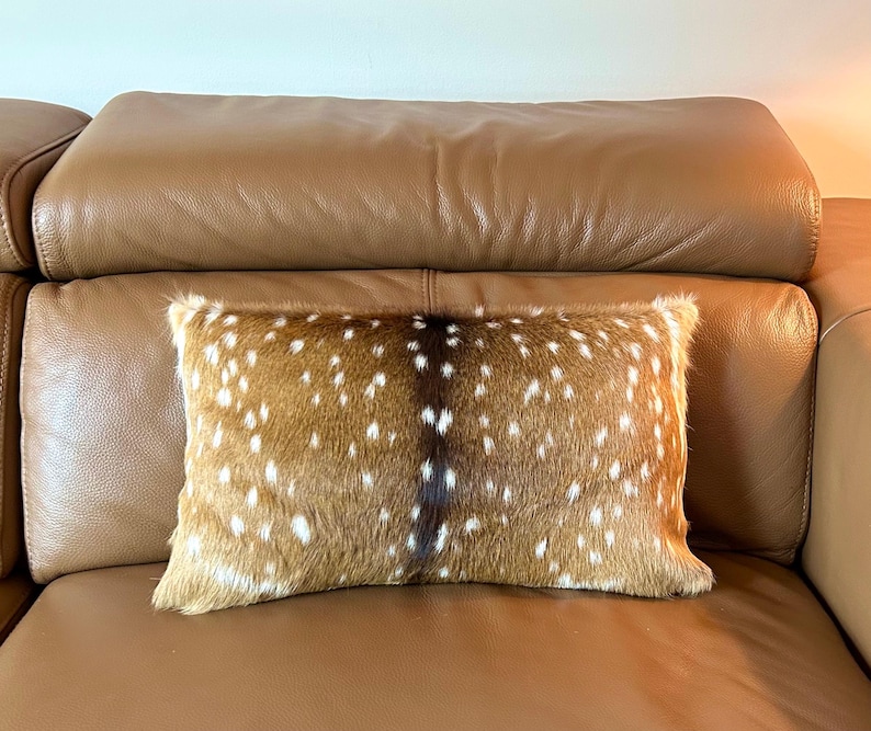 May include: A rectangular decorative pillow with a brown and white spotted pattern, resembling deer fur. The pillow is placed on a brown leather sofa, adding a touch of rustic charm to the interior decor.