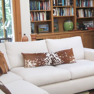 May include: A white sectional sofa with a brown and white cowhide patterned throw pillow and two square pillows. The sofa is in a living room with a bookshelf and a stone fireplace in the background.
